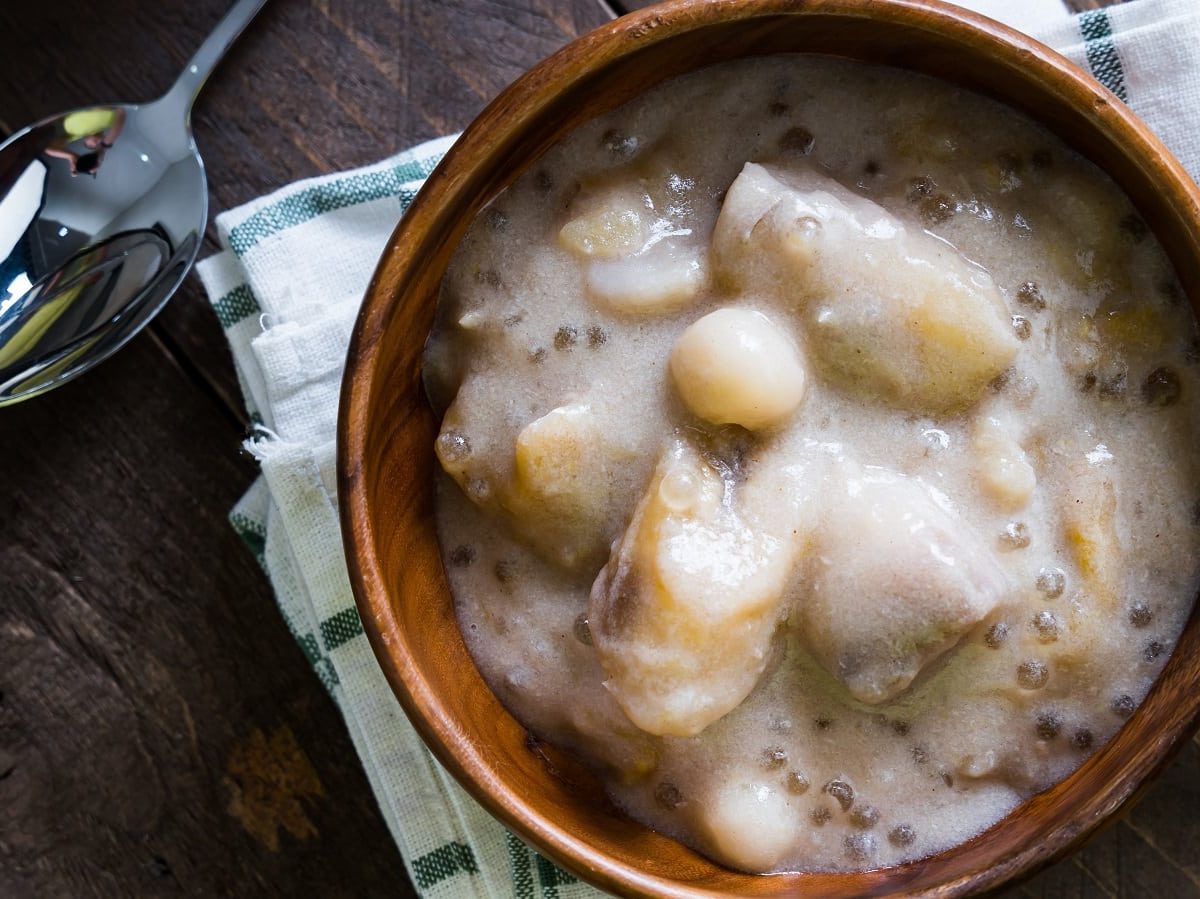 Eine Holzschüssel mit einer Portion der Sago-Suppe mit Taro und Bananen auf einem karierten Tischtuch neben einem Löffel, vor einem dunklen Holzhintergrund von oben.