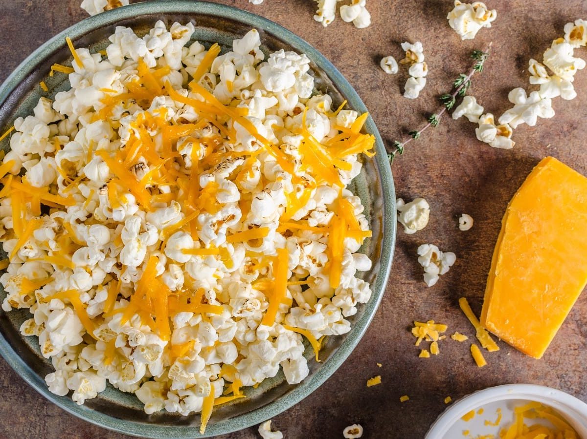 Eine große Schüssel gefüllt mit salzigem Popcorn mit Cheddar Streifen garniert, neben einzelnen Popcorn-Stücken, einem Thymian-Stängel und einem Stück Cheddar, vor einem braunem Hintergrund, von oben.