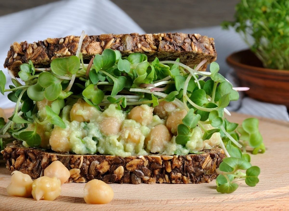 Ein Sandwich mit Kichererbsen-Guacamole und Sprossen auf einem runden Holzbrett. Um das Sandwich herum liegen einige weitere Kichererbsen.