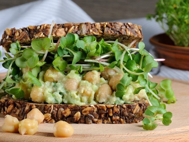 Ein Sandwich mit Kichererbsen-Guacamole und Sprossen auf einem runden Holzbrett. Um das Sandwich herum liegen einige weitere Kichererbsen.