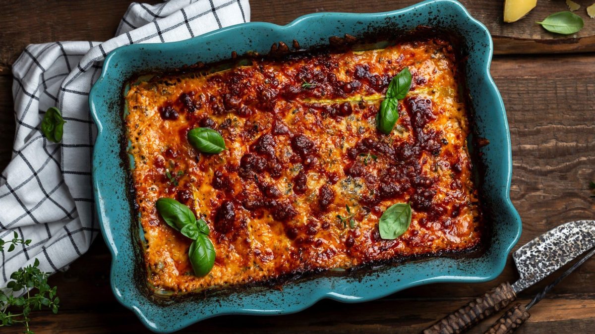 EIne blaue Auflaufform mit Sauerkraut-Lasagne und einigen Basilikumblättern in der Draufsicht, daneben Besteck und ein Geschirrtuch.