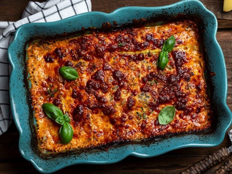 EIne blaue Auflaufform mit Sauerkraut-Lasagne und einigen Basilikumblättern in der Draufsicht, daneben Besteck und ein Geschirrtuch.