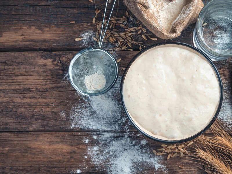 Eine Schüssel mit Sauerteig in der Draufsicht, darum herum Zutaten wie Mehl und Wasser, ein Sieb und Getreide.