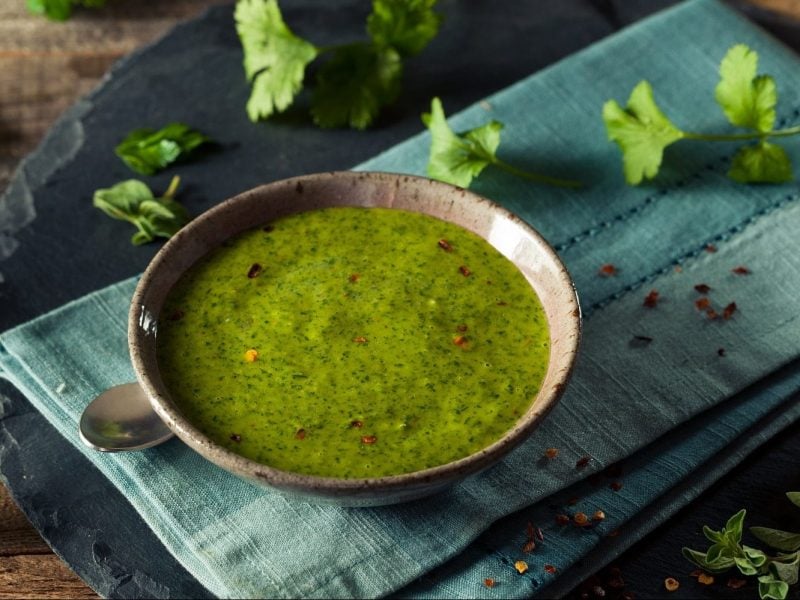 Auf einem türkisem Tuch steht eine Schale mit Salsa Verde. Neben der scharfen Sauce lieben Petersilienblätter.