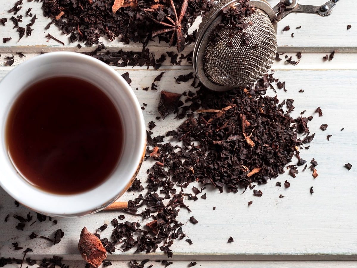 Eine weiße Tasse gefüllt mit dunklem Schwarztee neben einem Teesieb und losem Schwarztee der mit verschiedenen Gewürzen verfeinert ist.