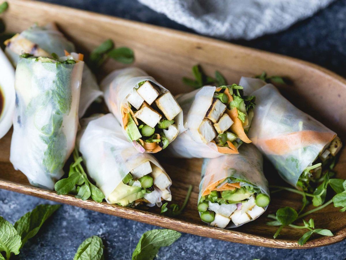 Selbst gemachte Sommer-Rollen, gefüllt mit mariniertem und geröstetem Tofu, Karotte, Spargel, Minze, Koriander und Zwiebeln, serviert mit malziger Sojasauce auf einem hölzernem Brett auf dunklem Untergrund