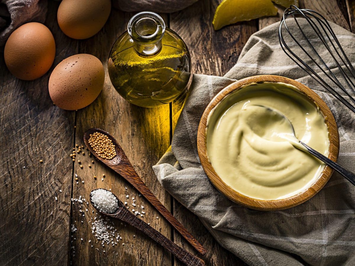 Auf einem Holztisch steht eine Holzschüssel mit selbstgemachter Mayonnaise. Neben ihr liegen ein Schneebesen, Eier, Öl, Salz und Pfeffer.