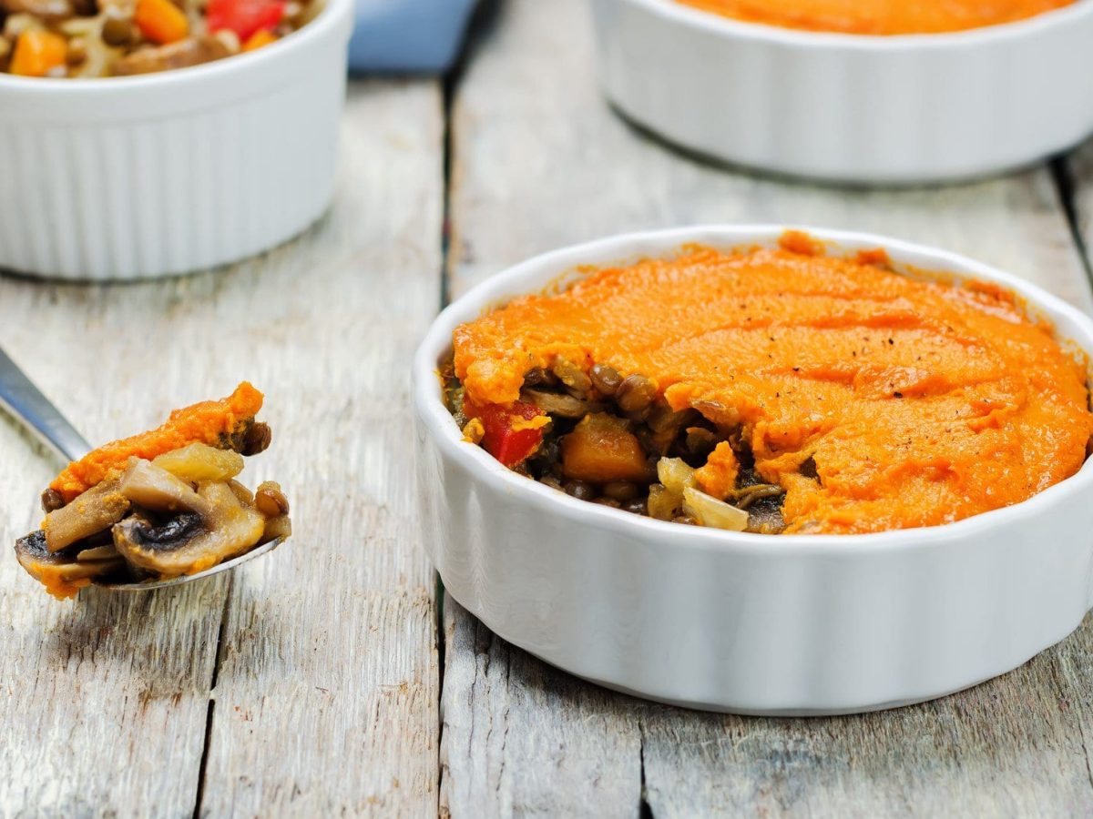 Eine runde Auflaufform Shepherd's Pie mit Süßkartoffelpüree, im Hintergrund sind noch zwei Formen zu erkennen, außerdem liegt ein Löffel mit gekochtem Gemüse daneben.