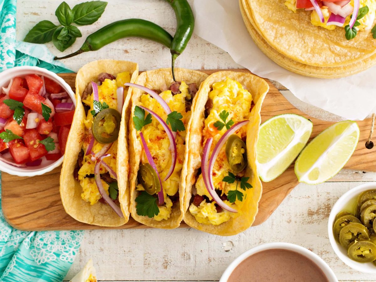 Drei Sin City Breakfast Tacos mit Omelette gefüllt auf Holzbrett . Daneben Tomatensalat. Recht Schälchen mit Jalapenos und Limettenspalten. Peperoni und Koriander als Deko im Bild verteilt. Aufnahme aus der Vogelperspektive.