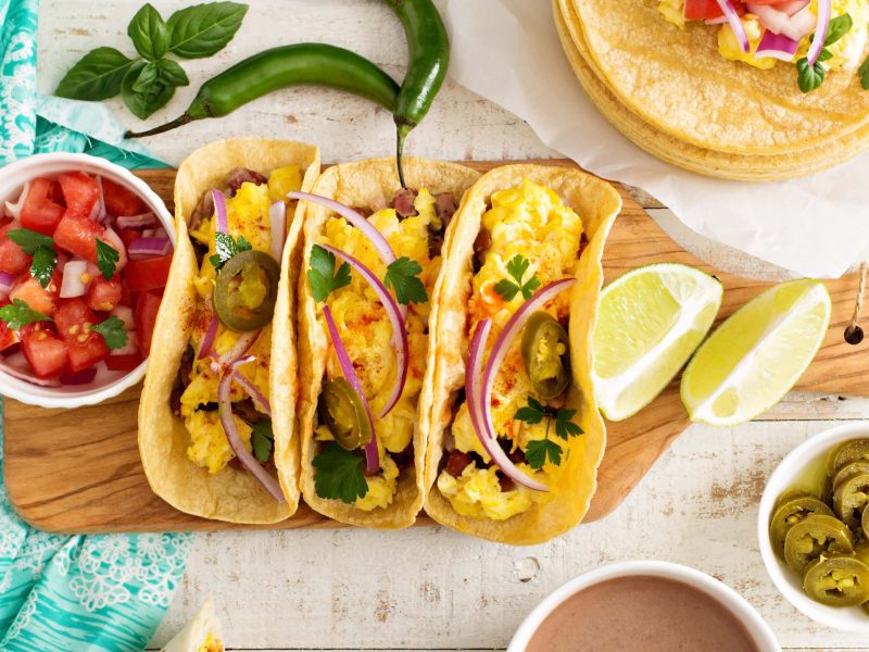 Drei Sin City Breakfast Tacos mit Omelette gefüllt auf Holzbrett . Daneben Tomatensalat. Recht Schälchen mit Jalapenos und Limettenspalten. Peperoni und Koriander als Deko im Bild verteilt. Aufnahme aus der Vogelperspektive.