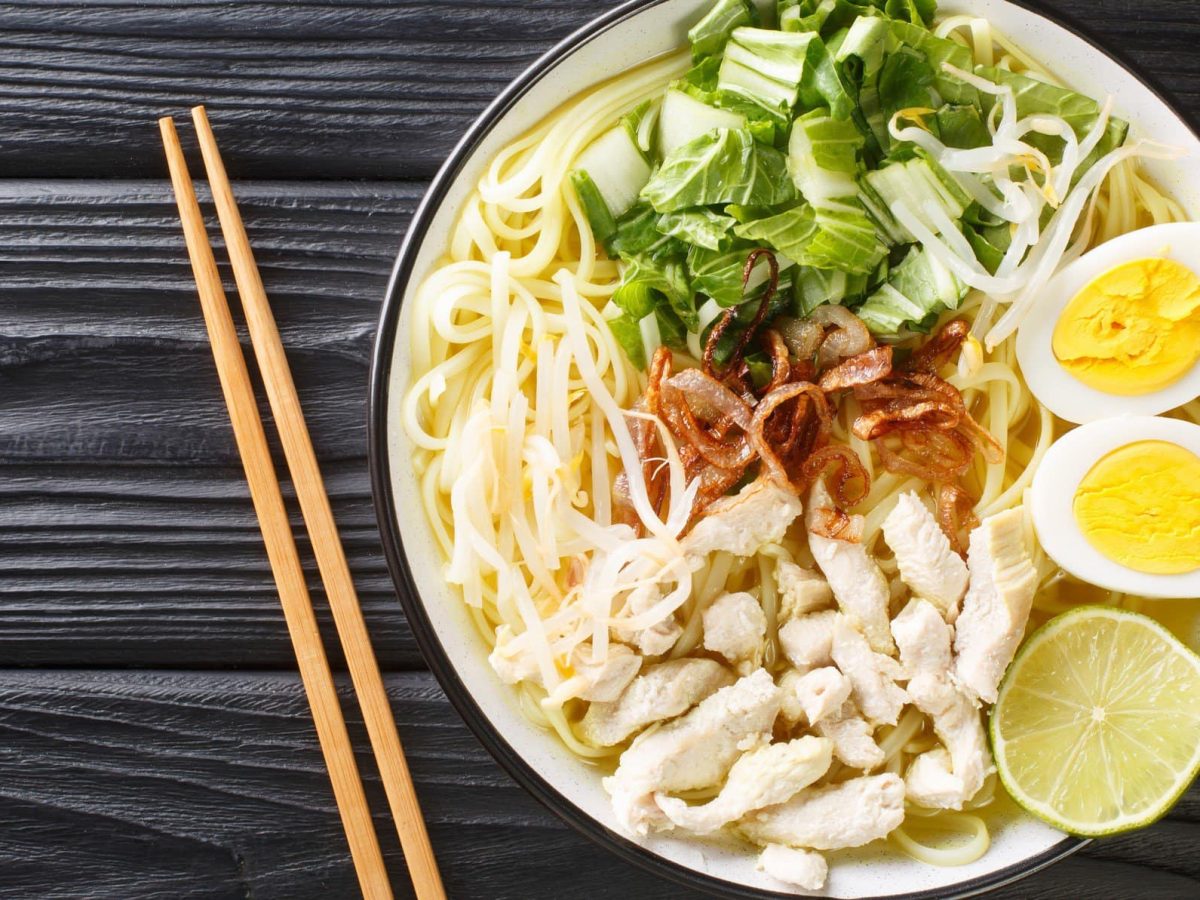 Soto Ayam indonesische Hühnersuppe mit Einlagen in weißer Suppenschüssel mit schwarzem Rand auf schwarzem Holztisch. Daneben Essstäbchen. Aufnahme aus der Vogelperspektive.