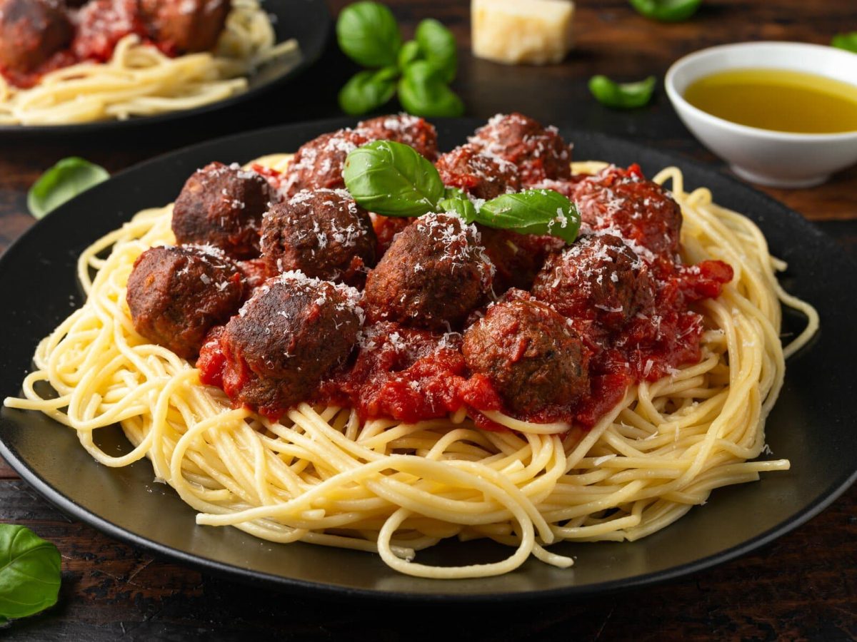 Ein Teller Spaghetti mit veganen Fleischbällchen, daneben liegen Basilikumblätter und ein paar Stücke Parmesan.