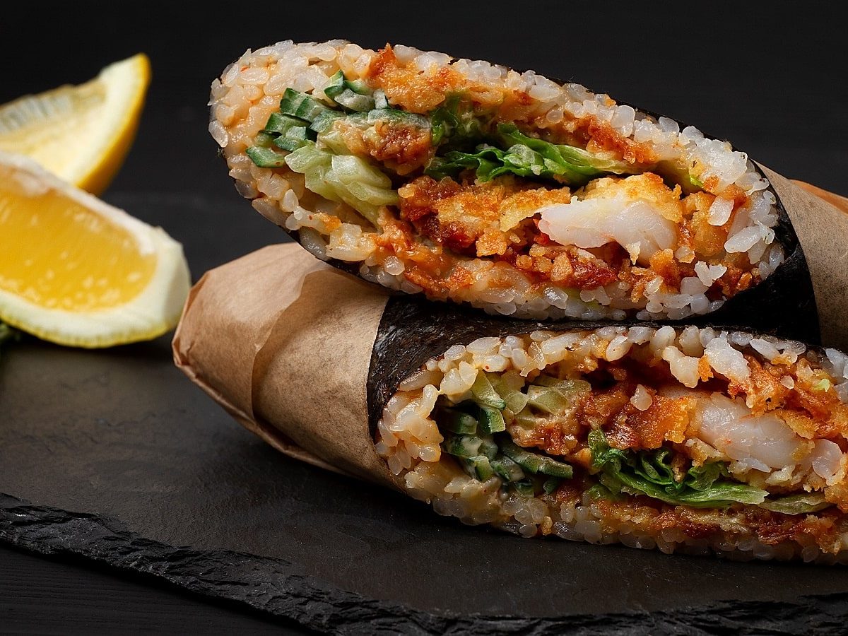 Ein aufgeschnittener Sushi-Burrito mit Shrimps-Tempura und scharfer Sauce in Papier eingewickelt, übereinander gelegen auf einer schwarzen Platte neben Zitronen und Kräutern vor einem schwarzem Hintergrund.