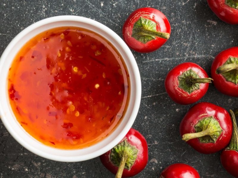 Eine weiße Schale Sweet Chili Sauce auf dunklem Untergrund in der Draufsicht, daneben liegen rote Chilischoten.