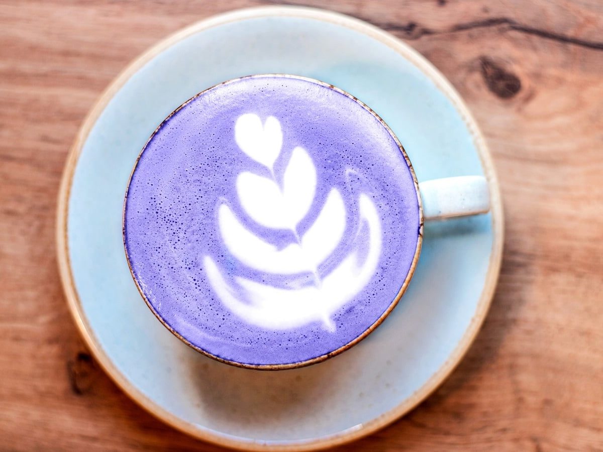 Eine Tasse Taro-Latte mit einer weißen Schaum-Verziehrung, auf einer Untertasse stehend, vor einem Holzhintergrund, von oben.