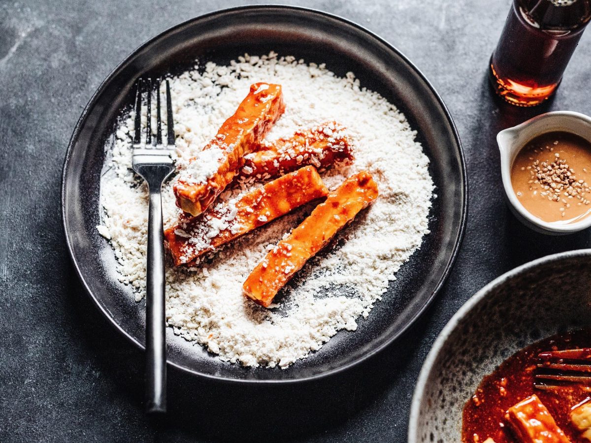 Tempeh-Fritten in der Zubereitung, mariniert und in Mehl gewendet, bereit frittiert zu werden. Daneben stehen Erdnusssauce und Fischsauce bereit, auf dunklem Hintergrund