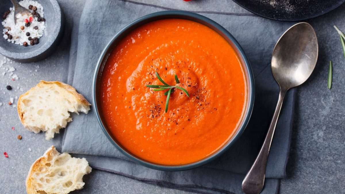 Tomaten-Erdnuss-Suppe in einer schwarzen Schale auf einem schwarzen Tuch, garniert mit Rosmarin und serviert mit frischem Weißbrot und grobem Salz und Pfeffer