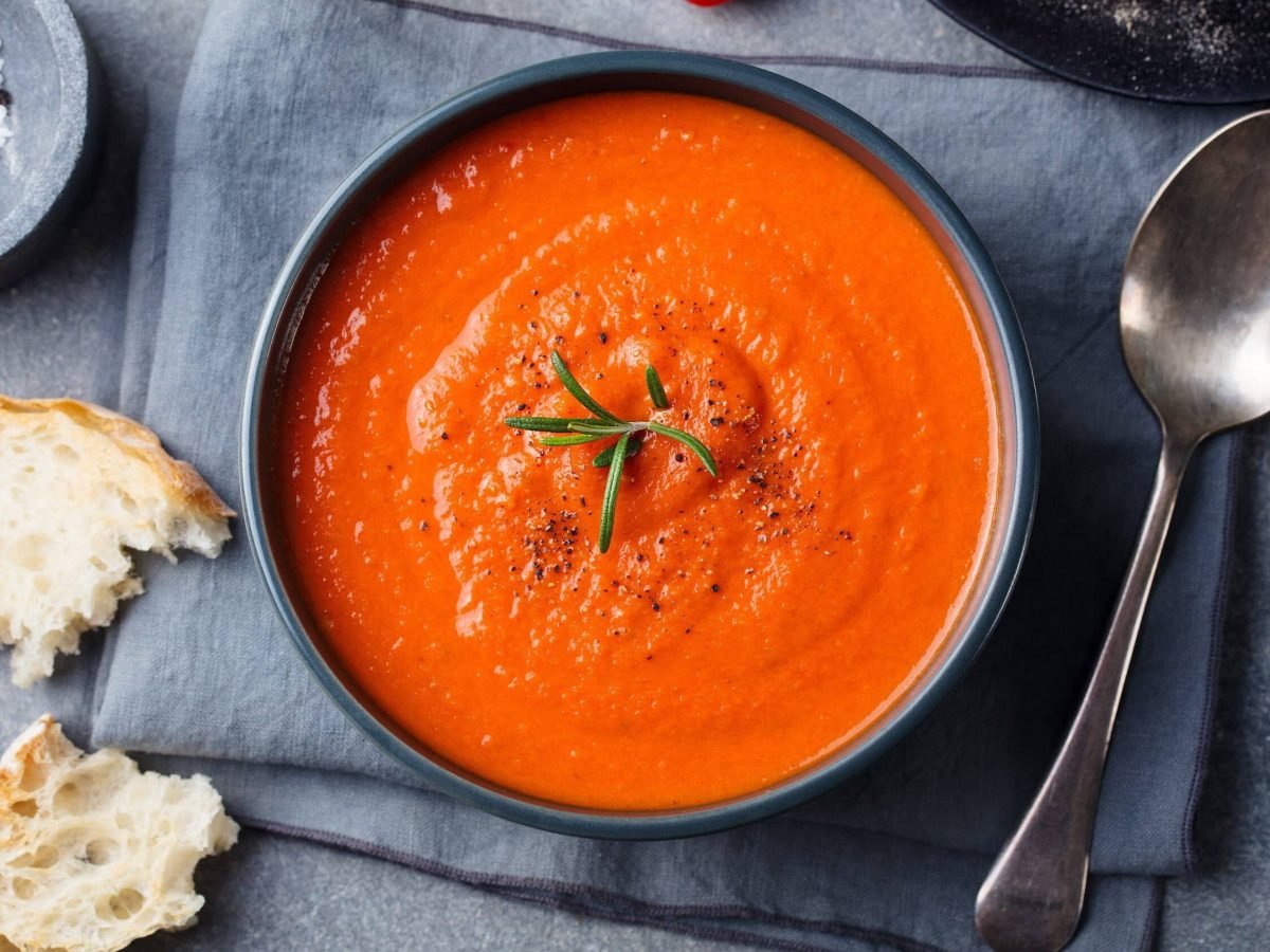 Tomaten-Erdnuss-Suppe in einer schwarzen Schale auf einem schwarzen Tuch, garniert mit Rosmarin und serviert mit frischem Weißbrot und grobem Salz und Pfeffer