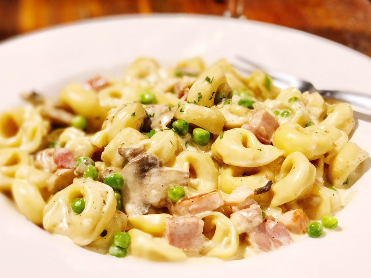 Tortellini à la creme aus Das große Fressen auf weißem Pastateller und dunkelbraunem Tisch. Rechts eine Gabel auf dem Teller. Aufnahme aus der Draufsicht.