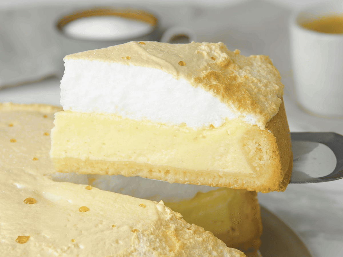 Ein Stück Tränenkuchen wird mit Hilfe eines Kuchenhebers von dem Rest des auf einer Kuchenplatte stehenden Tränenkuchens gehoben, sodass man die Haube, den Belag und den Boden von der Seite sehen kann. Im Hintergrund ist eine Tasse Kaffee sowie Zucker zu sehen vor einem grauen Hintergrund.