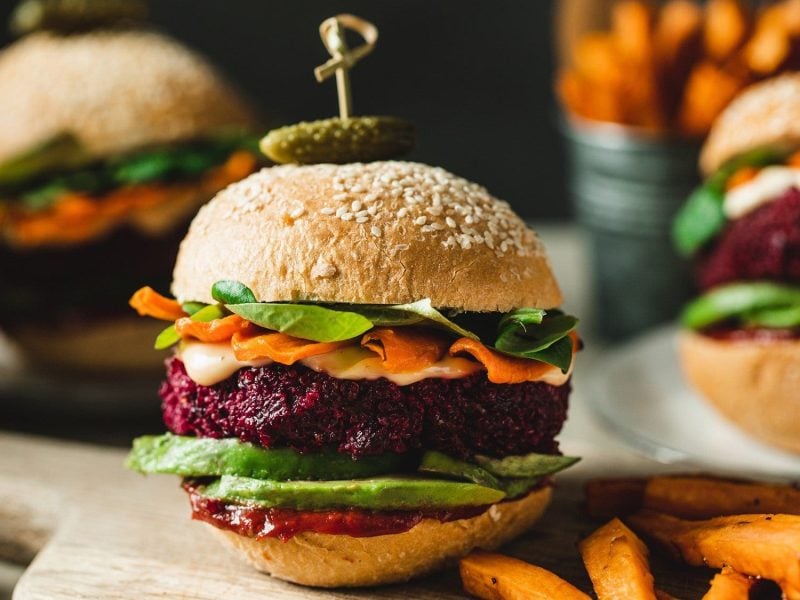 Vegane Burger mit Rote-Bete-Patty und Avocado liegen auf einem hellen Holzbrett. Die Burger sind zusätzlich angerichtet mit Mayonnaise, BBQ-Sauce, Karotten und Salat