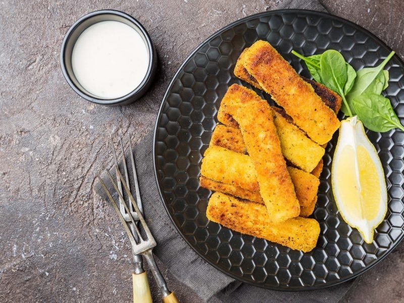 Vegane Fischstäbchen auf einem schwarzen Teller mit zitrone und Spinatblättern serviert. Daneben ein Schälchen mit Mayonnaise auf einem steinernen Untergrund.