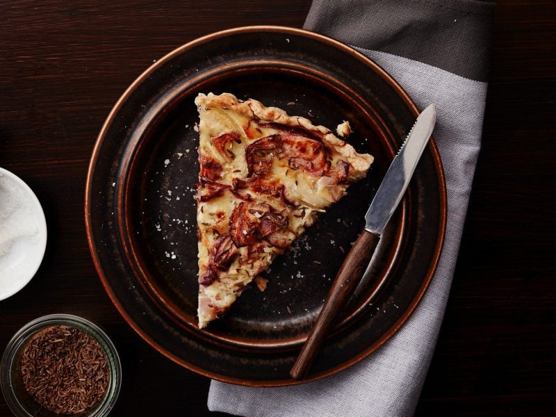 Ein Teller vegane Quiche Lorraine mit einem Messer auf dunklem Untergrund in der Draufsicht. Daneben eine Schale mit Salz und Kümmel.