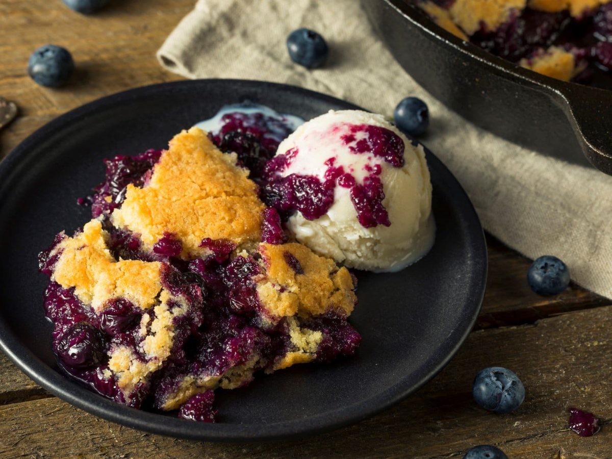 Ein Teller veganer Heidelbeerauflauf mit einer Kugel Vanilleeis, daneben liegen ein Löffel und einige Heidelbeeren verstreut.