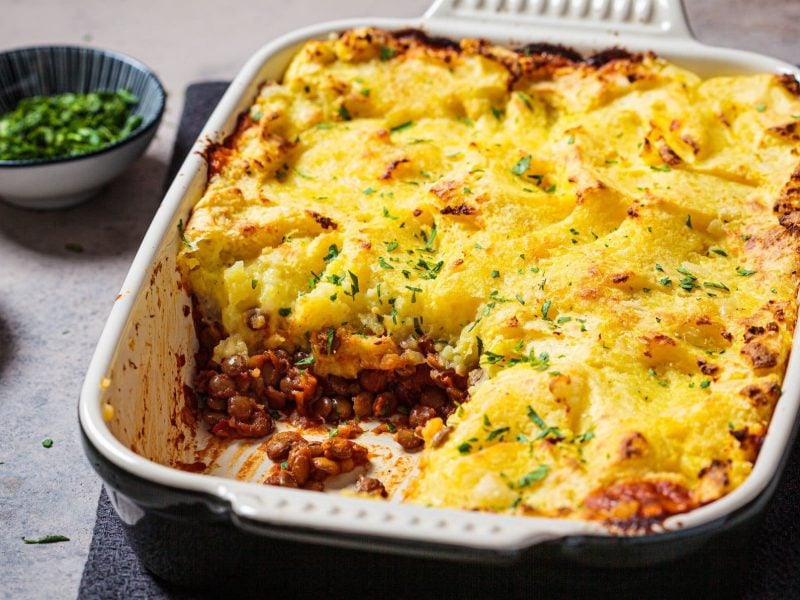 Eine Auflaufform mit veganem Shepherd's Pie, aus der schon ein Stück fehlt, daneben eine Schale mit Kräutern.