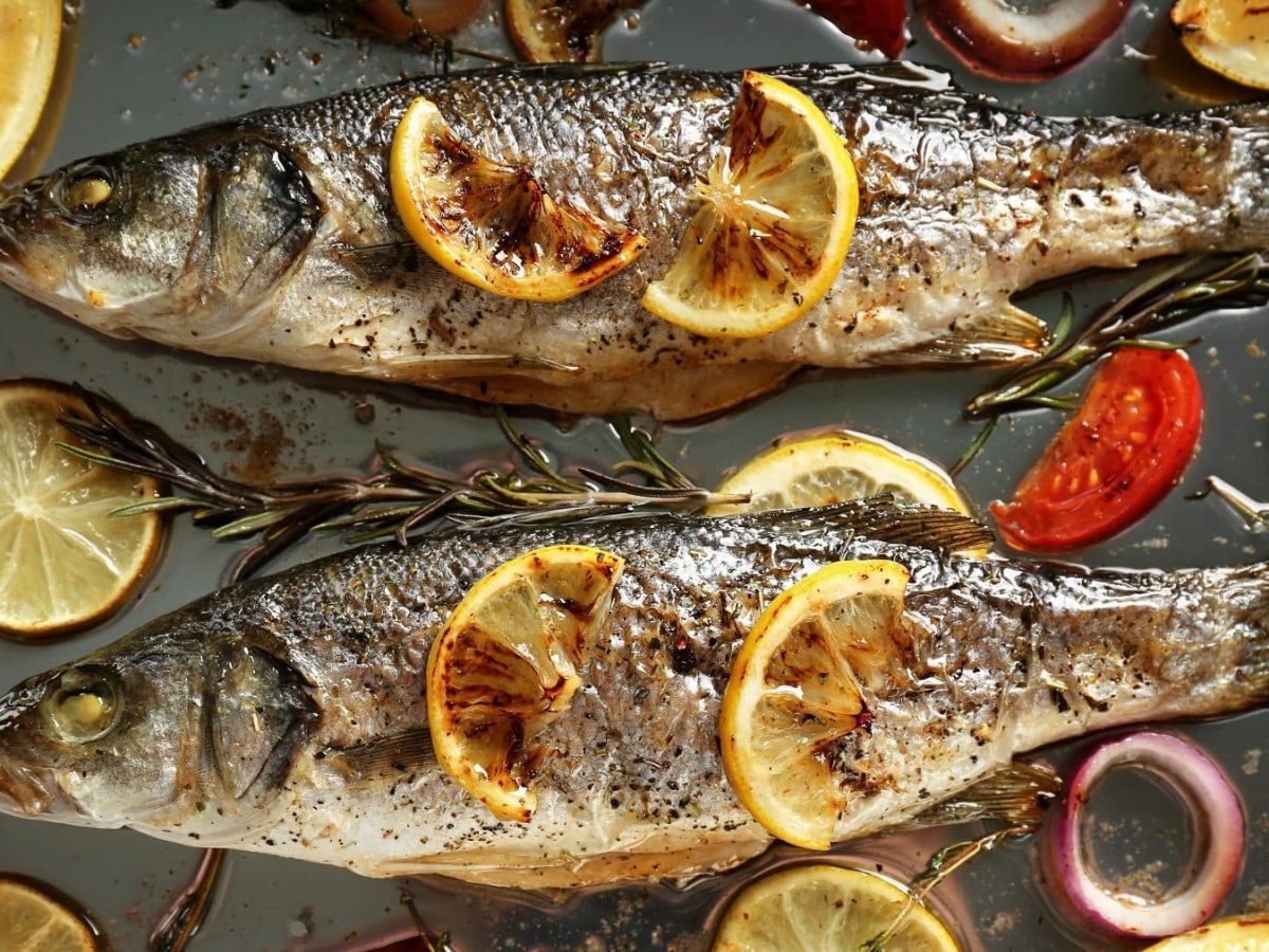 Aus der Vogelperspektive: Zwei Fische, es handelt sich um Wolfsbarsche, liegen auf einem Blech. Drum herum liegen Tomaten und Zitronenschieben sowie Rosmarin und Thymian.