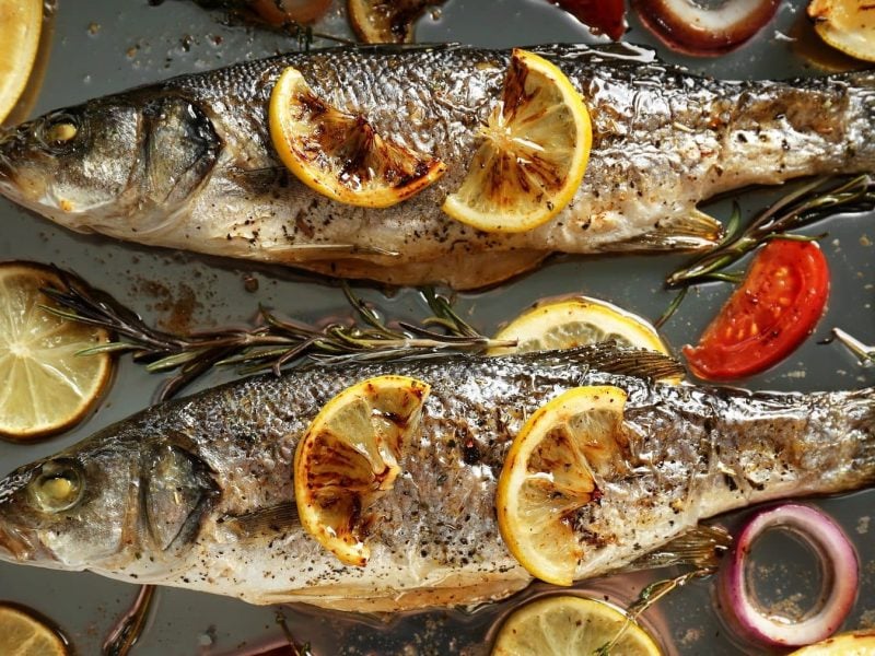 Aus der Vogelperspektive: Zwei Fische, es handelt sich um Wolfsbarsche, liegen auf einem Blech. Drum herum liegen Tomaten und Zitronenschieben sowie Rosmarin und Thymian.