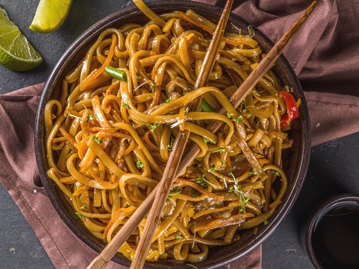 Eine dunkle Keramik Schale mit einer großen Portion der Yi Mein, der chinesischen Nudeln, die ein langes Leben bringen sollen, garniert mit Knoblauch-Schnittlauch und Chili, mit einem paar Essstäbchen, neben einer kleinen Schüssel der Sauce und zwei Limetten Scheiben, auf einem braunen Küchentuch, vor einem grauen Hintergrund, von oben.