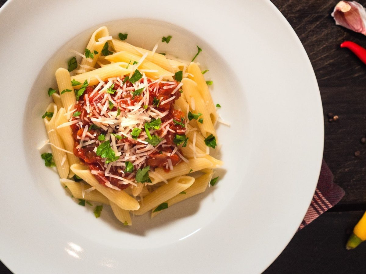 Penne all'arrabiata auf weißem Teller bestreut mit Pecorino und Petersilie auf dunklem Untergrund.