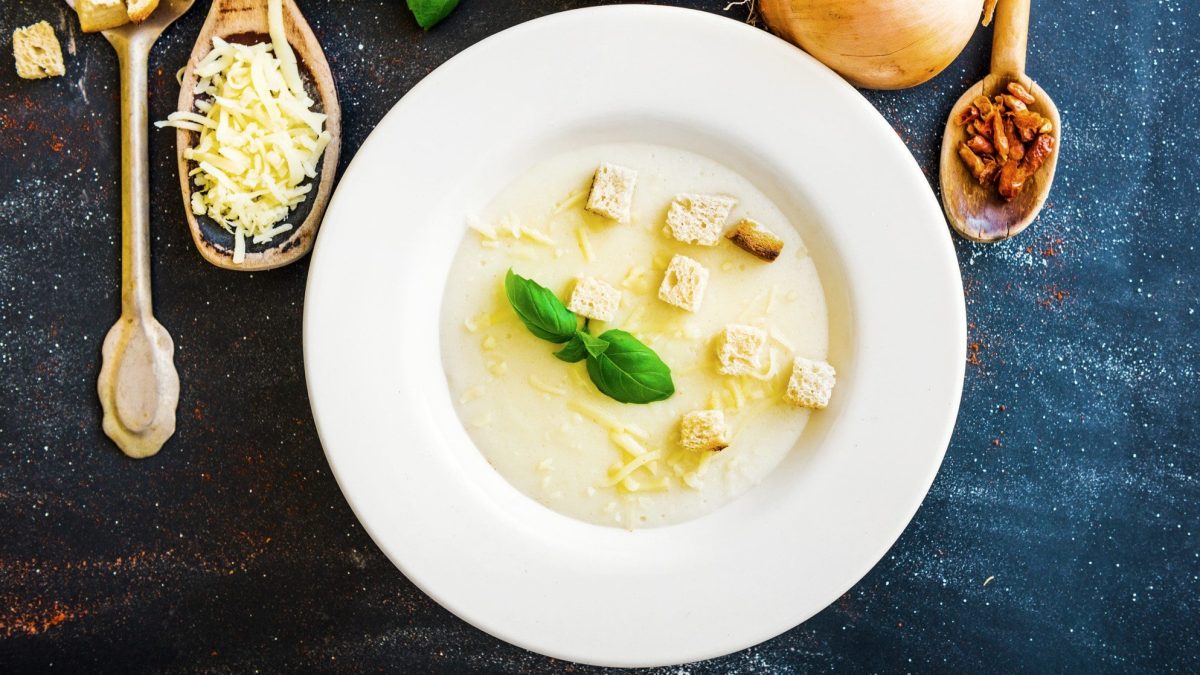 Weißer Teller mit Bergkäsesuppe und Croutons auf dunkelblauem Untergrund.