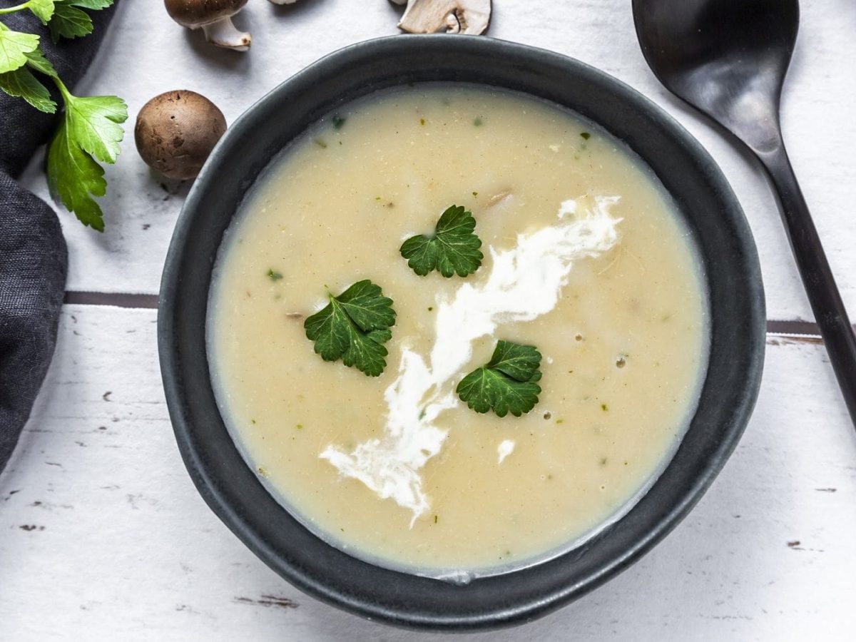 Aus der Vogelperspektive: Auf einem Tisch steht eine cremige Champignonsuppe in einer schwarzen Schale. Daneben liegen Persilienzweige, ein Löffel und einige Scheiben Weißbrot.