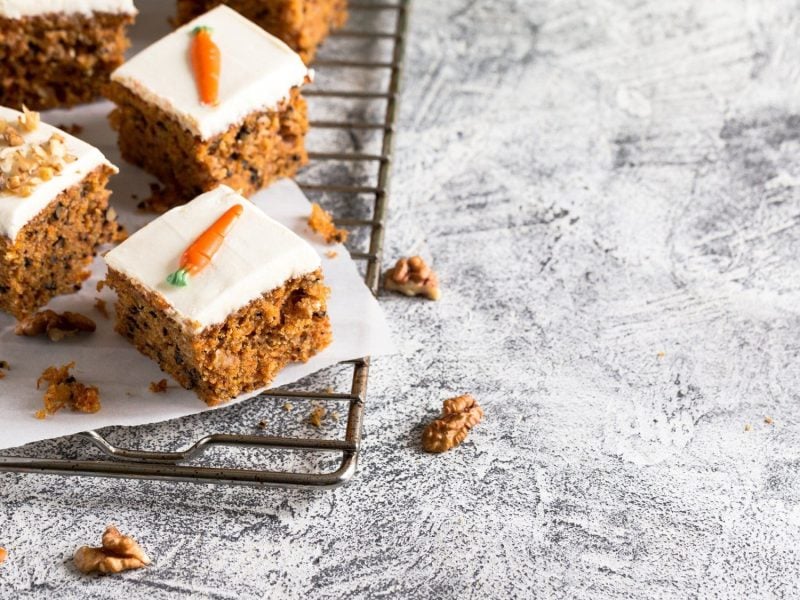 Auf einem Backgitter liegen einige Stück Karottenkuchen in Quadrate geschnitten. Sie sind dekoriert mit Frischkäse, Walnüssen und Marzipan-Karotten.