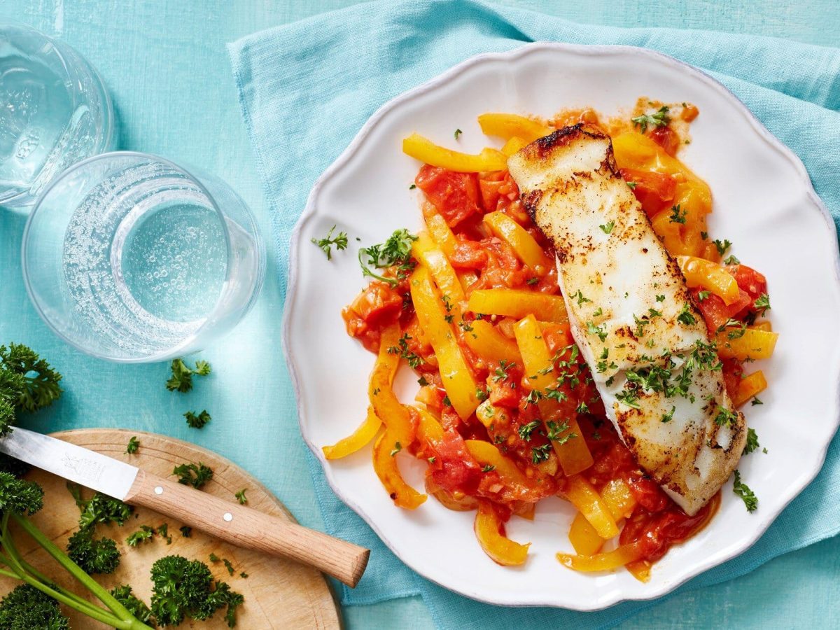 Fischfilet auf Paprikagemüse in Draufsicht mit Glas Petersilie und blauer Tischdecke