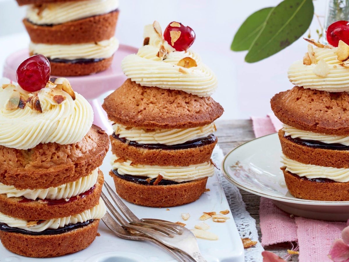 Auf einem Tisch sind 3 Frankfurter-Kranz-Cupcakes angerichtet. Sie bestehen aus drei Schichten mit Buttercreme und Kuvertüre. Obendrauf ist ein Tüpfel Buttercreme und jeweils eine Kirsche.