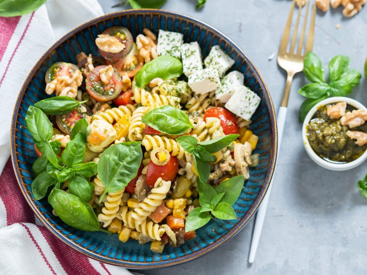 Unser italienischer Pasta Salat angerichtet in einer Schüssel. Im Hintergrund sind Tomaten, Pesto und Walnüsse zu sehen.