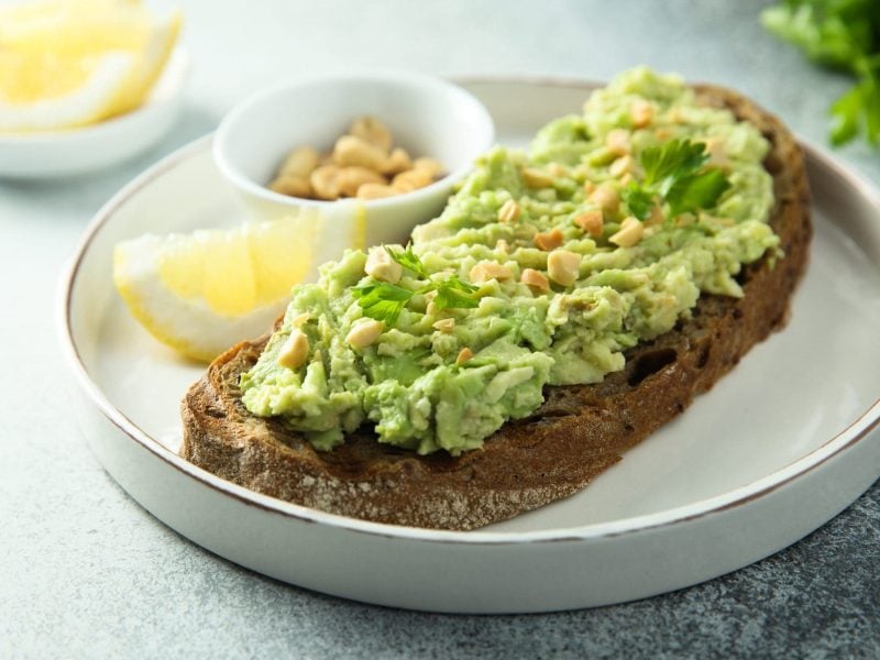 Unser Guacamole-Brot liegt angerichtet mit Erdnüssen und Zitronenschnitzen auf einem weißen Teller.