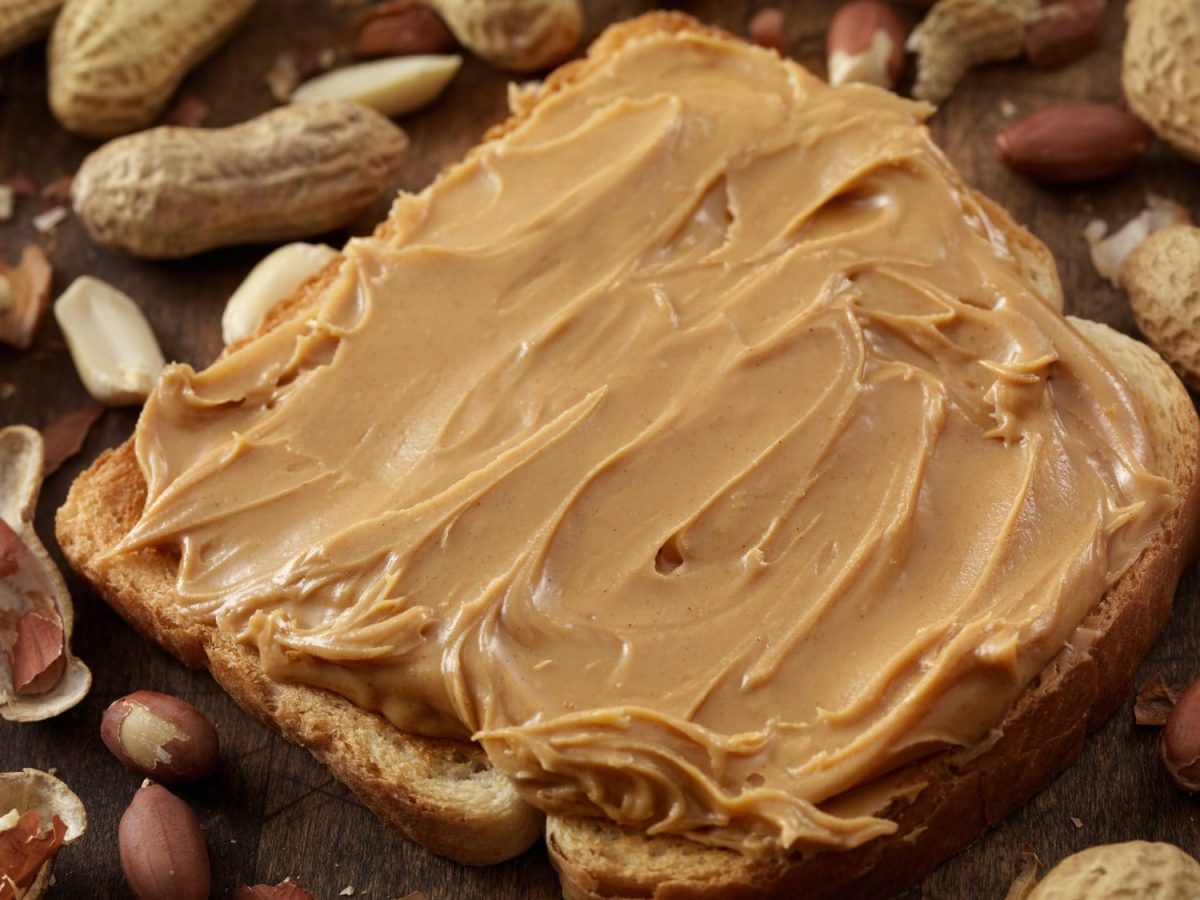 Eine Scheibe Erdnussbutter-Toast liegt inmitten von zahlreichen Erdnüssen auf dem Tisch.