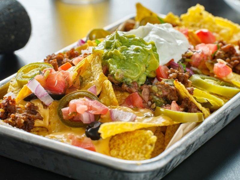 Loaded Nachos mit Chili in weißer Auflaufform auf grauem Untergrund. Links angedeutet dunkelgrauer Mörser und Olivenöl in Karaffe. Frontalansicht.