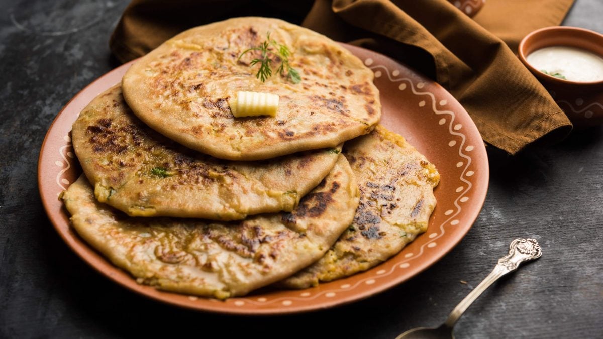 Aloo Paratha auf einem braunen Teller. Daneben ein Küchentuch, ein Löffel und ein Dip. Das Ganze auf einem hölzernen Untergrund.