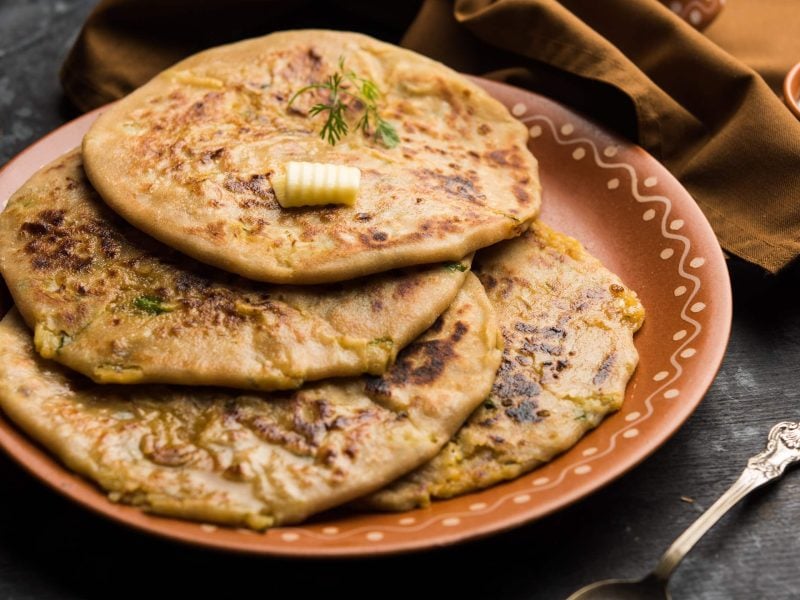 Aloo Paratha auf einem braunen Teller. Daneben ein Küchentuch, ein Löffel und ein Dip. Das Ganze auf einem hölzernen Untergrund.