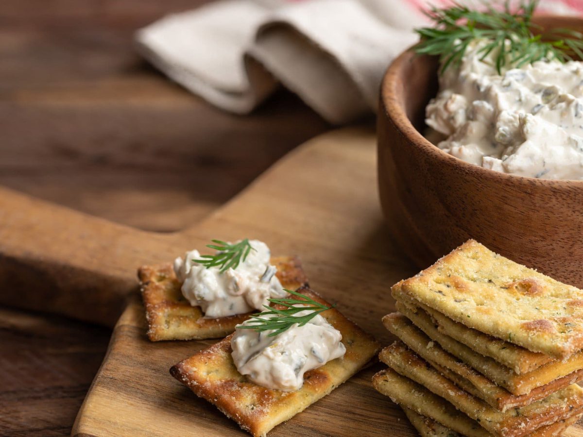 Bacon Onion Dip in einer Tonschale mit Crackern auf Holzbrett auf Holztisch. Weißes Tuch im Hintergrund. Frontalansicht.