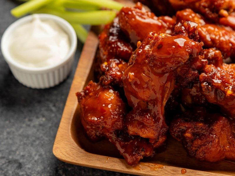 Blazin' Buffalo Wings auf Holzplatte angerichtet. Kleine Schale mit Creme Fraiche und Selleriestangen im Hintergrund. Platziert auf grauem Untergrund. Frontalansicht.