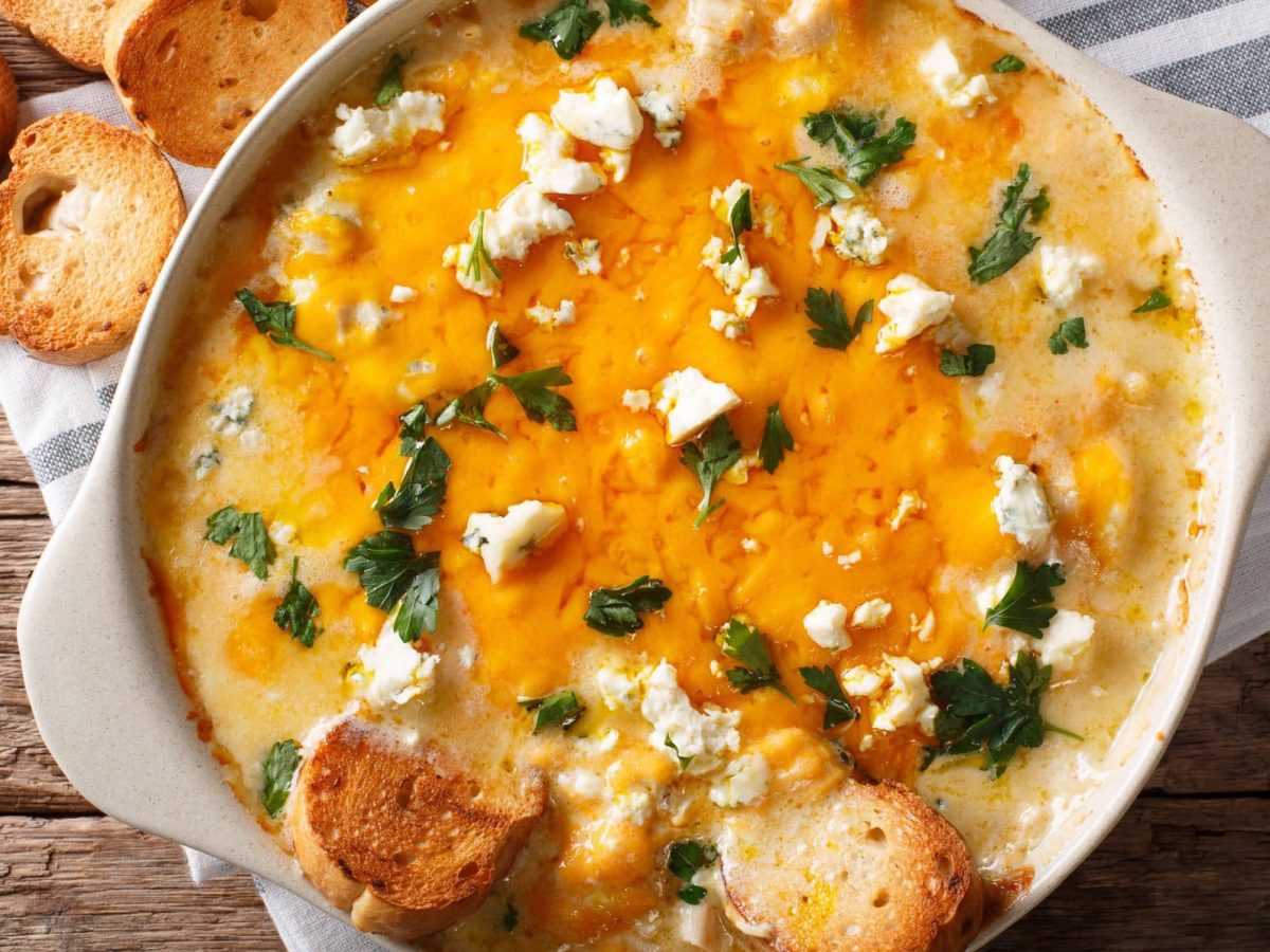 Buffalo Blue Cheese Dip in weißer Ofenform auf Tuch auf Holztisch. Auf dem Dip und daneben geröstete Brotscheiben. Aufnahme aus der Vogelperspektive.