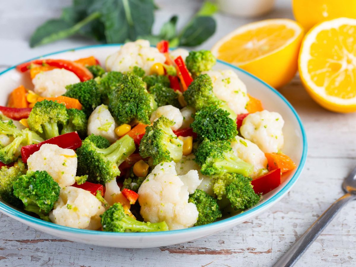 Ein Teller Blumenkohl-Brokkoli-Salat mit Paprika und Möhren, daneben eine Gabel und halbierte Apfelsinen.