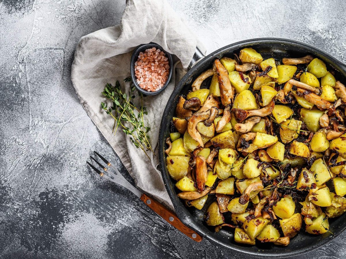 Rustikale gusseiserne Pfanne mit Bratkartoffeln mit Austernpilzen, garniert mit frischem Thymian auf einem bemehlten schwarzen Untergrund, von oben