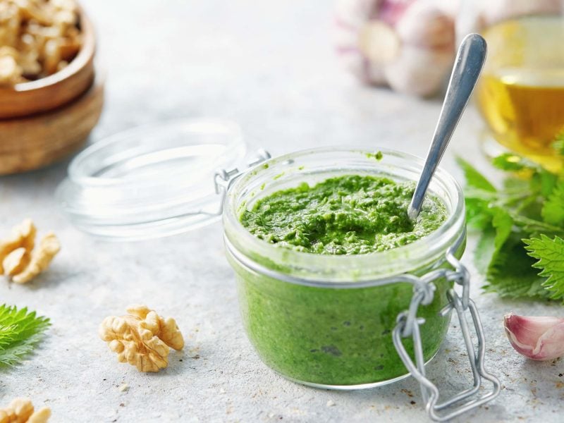 Ein Glas mit Brennnessel-Pesto mit Walnüssen. Im Huntergrund und daneben verschiedene Zutaten.